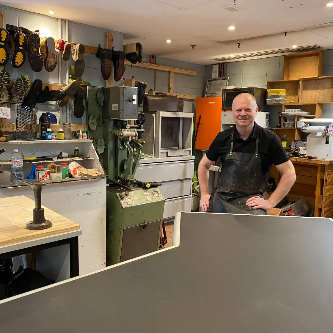 John Coburn in the workshop beside the green press, resoled boots on the rack behind him