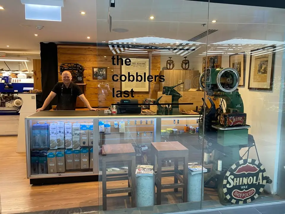 The shopfront in daylight — John behind the counter with shoe care products and the green stitcher